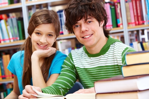 teen boy and girl study in library and smile for camera. The girl rests her chin in her palm.