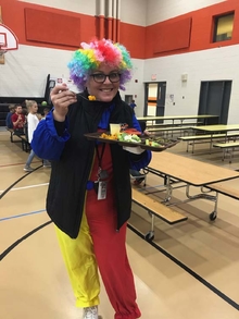 The principal of Menagha elementary wears a silly clown costume and is holding a tray of vegetables in the school cafeteria. 