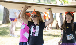 A group of girl campers carrying a large piece of cardboard over their heads.