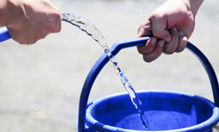 One hand filling a bucket with a water hose while another hand holds the bucket.