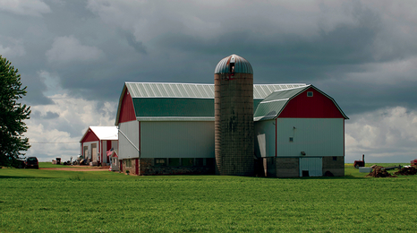 White and red barn with silo on a green field under a cloudy sky.