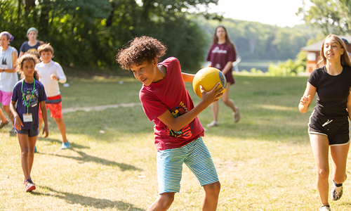 A boy holding a yellow ball outdoors while youth run around him.