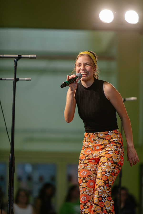 A teen girl wearing a 1970s outfit singing into a microphone on a stage.