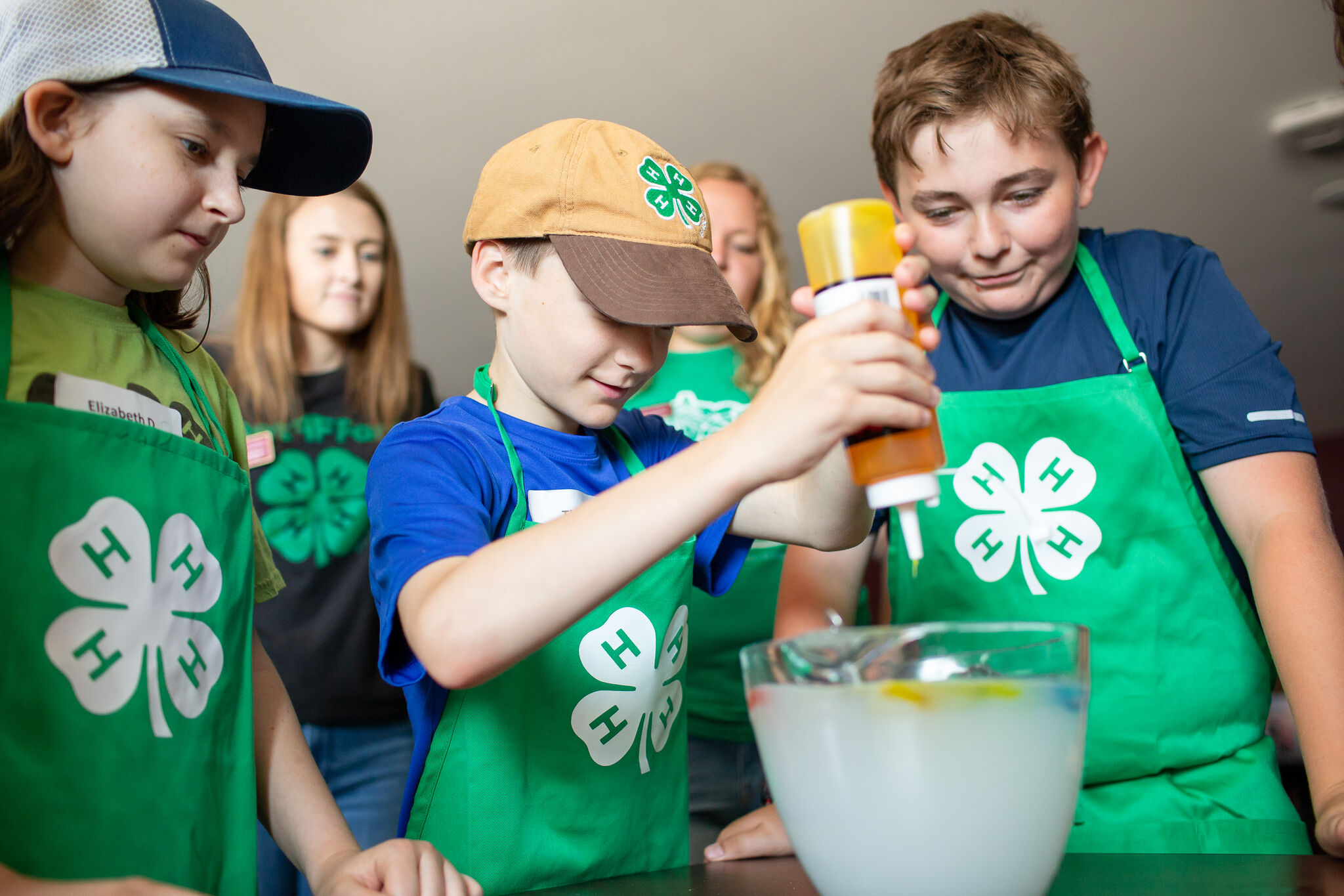 A group of youth wearing 4-H aprons with one boy putting sauce in a mixing bowl.