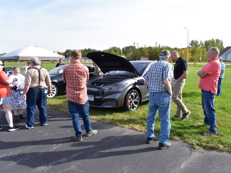 Group of people gathered around an electric vehicle with its hood up.