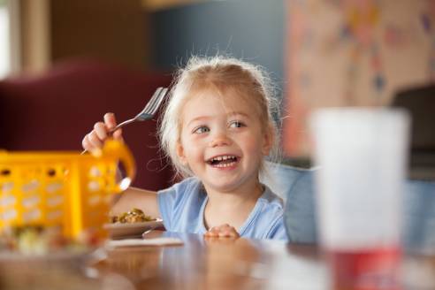 young girl at a table with food on a plate and a fork in her raised hand