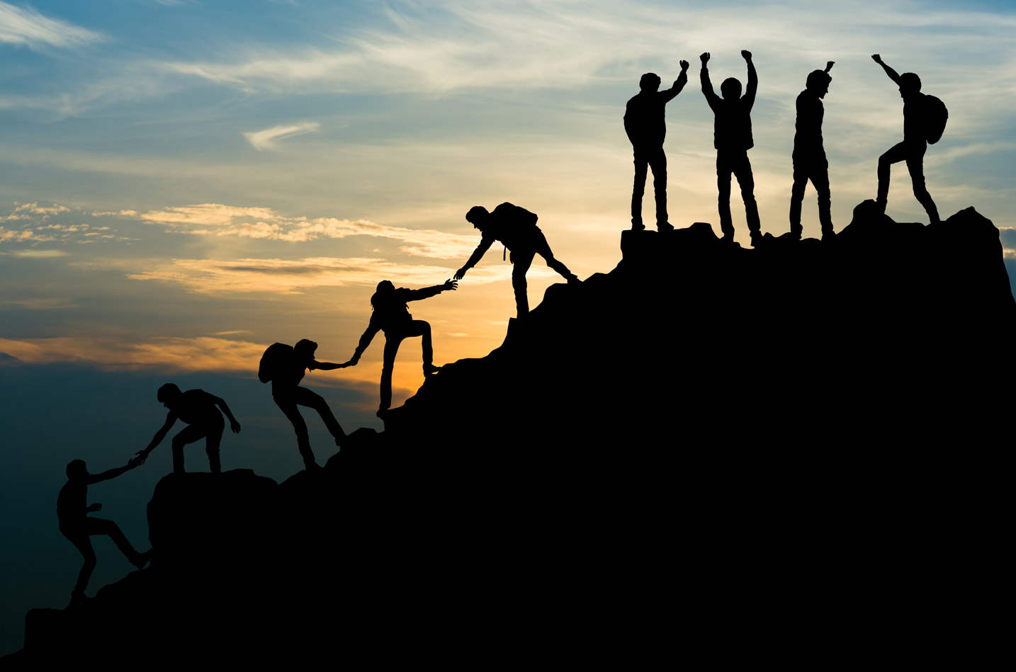 A silhouette of a group of people leading and helping each other climb a mountain as the sun sets.