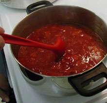 A red spoon stirring strawberry jam in a large metal pot.