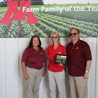 Extension's dean stands next to the farm couple holding their award plaque. A large "M" and "Farm Family of the Year" is on a wall behind them.