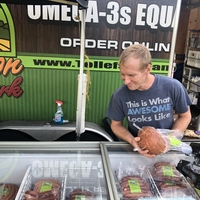 Man selling a ham out of a refrigerator case at farmers market wears shirt that says "This is what awesome looks like" and truck behind him says "Omega 3s"