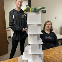Two girls next to a notecard tower with a stuffed animal bear lying on top of the tower.