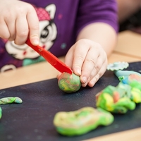 A close up have a child's hands cutting into green play doh with a red plastic knife. 