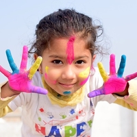 A girl with paint on her face has her hands up showing her painted palms.