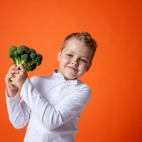 A boy wearing a white shirt holding a head of broccoli with a bright orange background behind him.