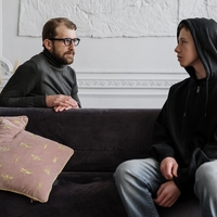A man talking to a teen boy wearing a hoodie sweatshirt sitting on a couch.