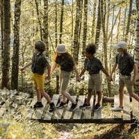 A line of youth holding hands while crossing a bridge in a wooded area.