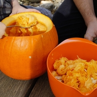 A close up of hands scooping out a pumpkin next to an orange bowl filled with the pumpkin's guts.