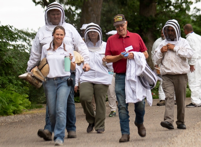 A group dressed in beekeeping apparel walks on a gravel road. The man in the middle holds his bee suit over his arm. He is wearing a Vietnam Veteran billed cap.