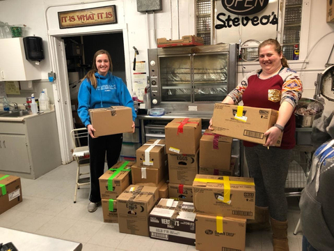 Two women hold grocery boxes smiling in a warehouse. They have packed 14-day meal kits stacked in front of them. 