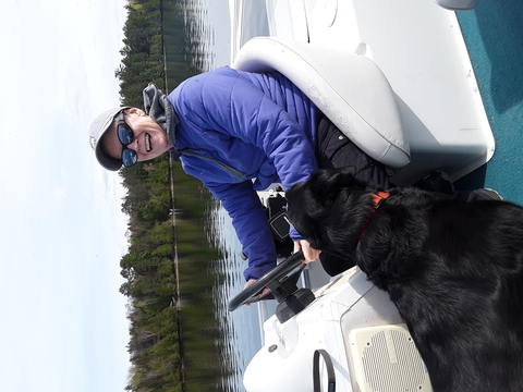 Woman drives a boat on a lake and her black dog is at her side.