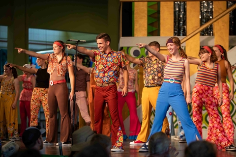 Youth on stage pointing, smiling, and wearing 1970s colorful stripes and floral prints