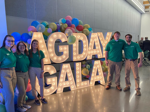Five youth wearing green 4-H polos standing in front of a large light up sign that reads, "Ag Day Gala."