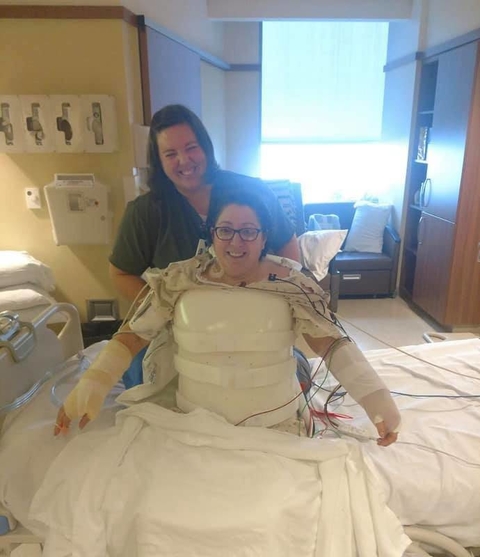 Anna in a hard plastic shell around her torso, casts on her arms while sitting on a hospital bed and a woman standing behind her supporting her.