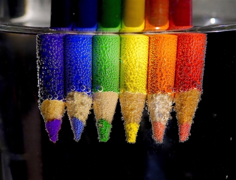 Photo of red, orange, yellow, green, and blue colored pencils that are submerged in water and are covered in small bubbles.