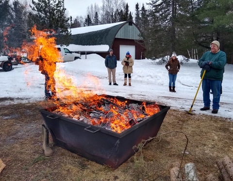 Adults standing behind a kiln of burning wood making biochar