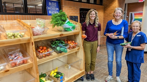 Three adults stand next to shelves of veggies, and each one is also holding a tomato, carrots, or a zucchini.