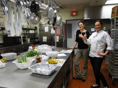 A chef and her assistant stand smiling with thumbs up in a commercial kitchen. Produce is on the counter in preparation for class. Pots, pans and other kitchen utensils hang from the ceiling. The kitchen countertop has fresh produce and recipes in preparation for cooking in the class.