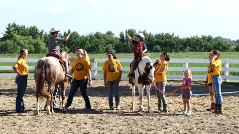 Two horses with kids riding wearing helmets as a group of youth and adults look on.