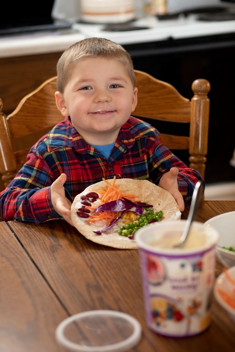 Preschooler boy in flannel shirt shows off his tortilla filled with peas, carrots and purple cabbage. He has a yogurt mustache.