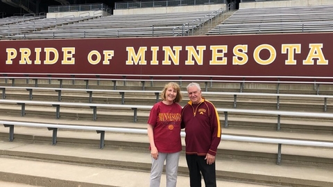 Couple stands in front of bleachers with "Pride of Minnesota" on a banner behind them