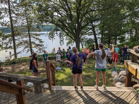 Two teens leading campers in song by a lake. 