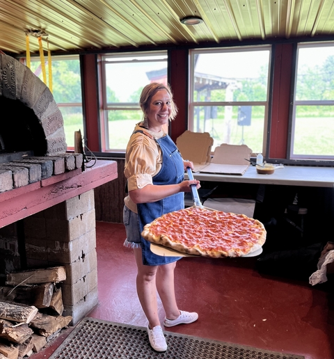 Chef Emily stands in front of her brick oven, which has logs below it, holding out a very large pepperoni pizza.