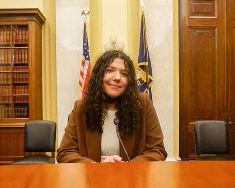 Teen sits at a table in the U.S. Capitol with two flags, a bookcase, and a door behind her.