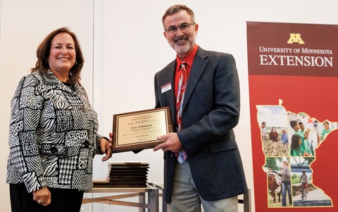 The dean and Lee Johnston stand together in front of Extension sign while Lee holds award plaque