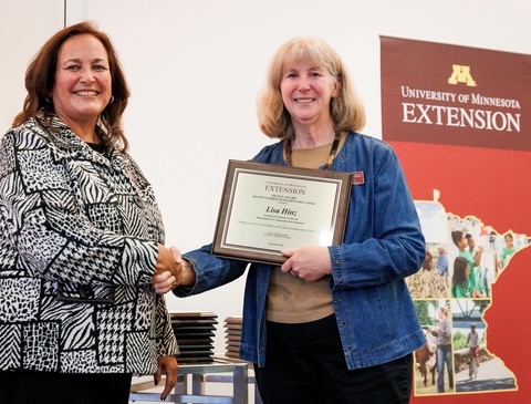 The dean and Lisa stand together in front of Extension sign while Lisa holds award plaque