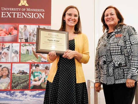 The dean and Natalie Hoidal stand together in front of Extension sign while Natalie holds award plaque