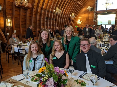 Three youth sitting at a fancy table with official 4-H name tags on and two youth members wearing green blazer standing behind them.