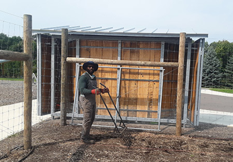 Man holding large metal broadfork in a bare garden bed with parking lot and shed behind. Garden is surrounded by a fence.