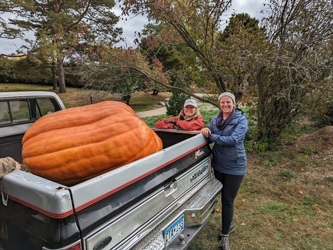 Giant orange pumpkin in the bed of a silver pickup with two women posing beside the truck.