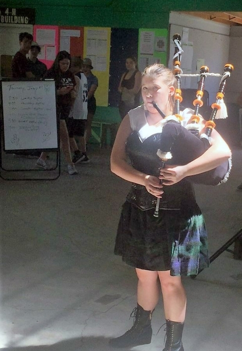 Victoria plays the bagpipes inside a building with light streaming through a window landing on her face. She's wearing a kilt and black work boots.