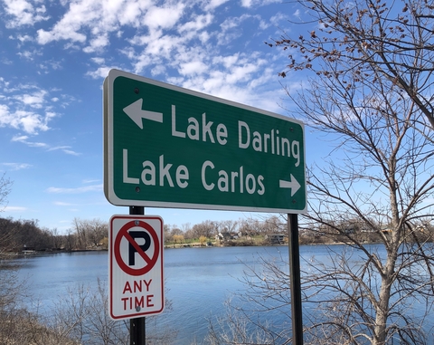 Sign next to lake pointing to Lake Carlos and also to Lake Darling in the other direction. There is also a no-parking sign. Sky is blue with a few white clouds.