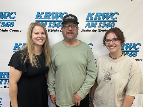 radio host poses between Emily Hansen and Emma Bruder with KRWC call sign on wall