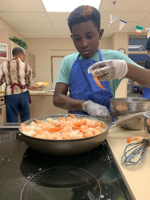 A boy wearing a kitchen apron focuses on his shrimp alfredo on the stove and a woman behind him is holding a plate