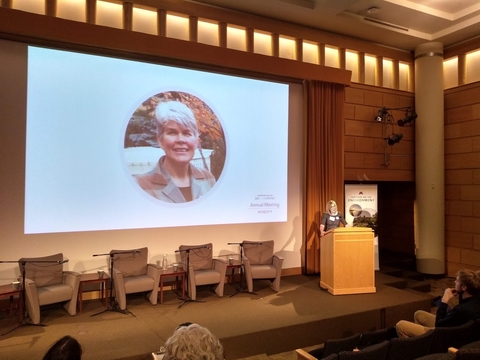 A woman standing behind a podium on a stage with a projector screen behind her displaying the face of former NWRSDP Executive Director, Linda Kingery.