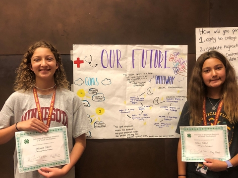 Two sisters stand on either sign of their "Our future" poster, holding certificates of completion for 4-H campus immersion.