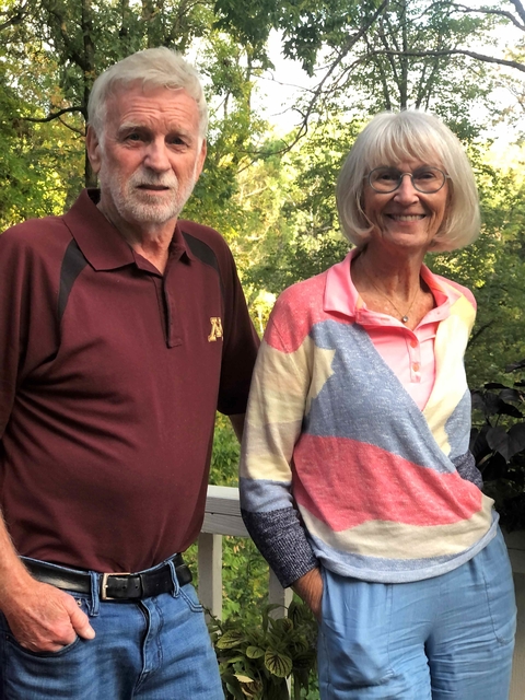 Married couple stand outdoors on a deck with trees behind them. They each have their right hand in their pocket.